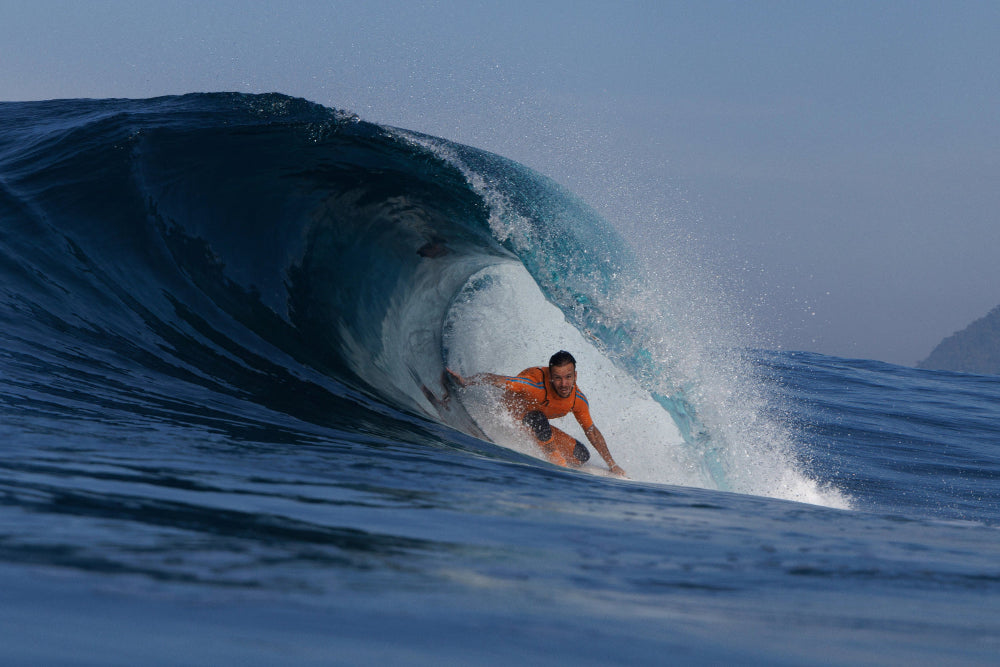 Surfer riding a large wave in the ocean with clear blue sky wearing Orange JANGA impact wetsuit