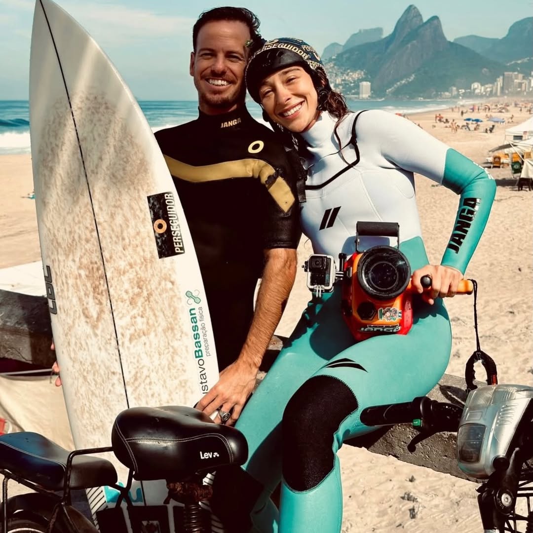 Smiling couple in wetsuits holding a surfboard and camera sitting by the beach with ocean and mountains in the background