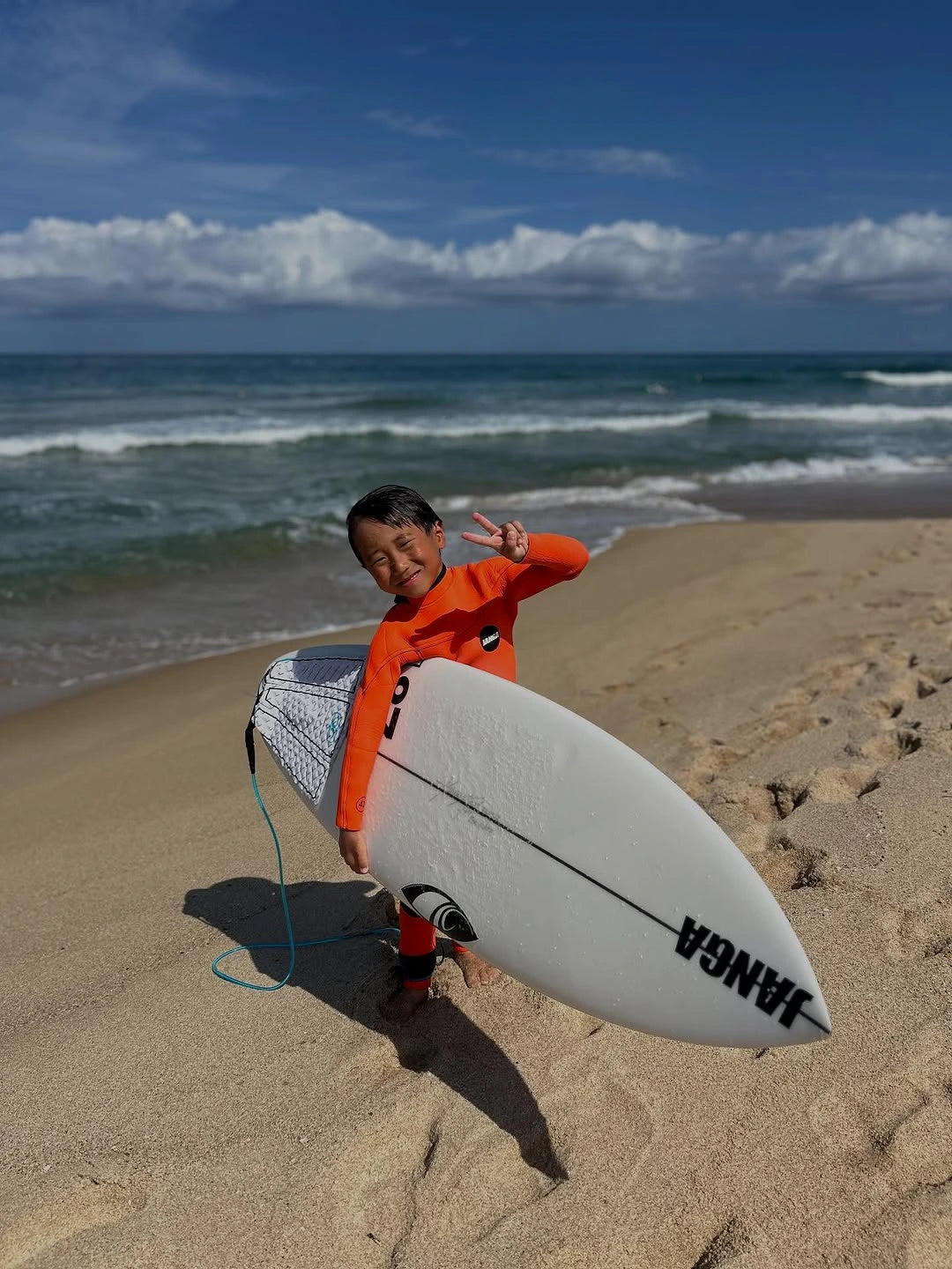 Child in an orange wetsuit holding a surfboard on a sandy beach with ocean and sky in the background